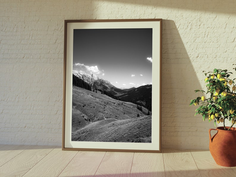 Op de afbeelding: Een zwart-witfoto van een bergketen met een bewolkte lucht. De foto is ingelijst in een bruin houten lijst en hangt aan een witte bakstenen muur.