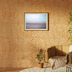 Op de afbeelding: Een bruine leren fauteuil met een wit plaid staat voor een bakstenen muur. Een ingelijst beeld van een strandsc&egrave;ne hangt aan de muur boven de stoel. Een kleine plant in een pot staat op de vloer naast de stoel.