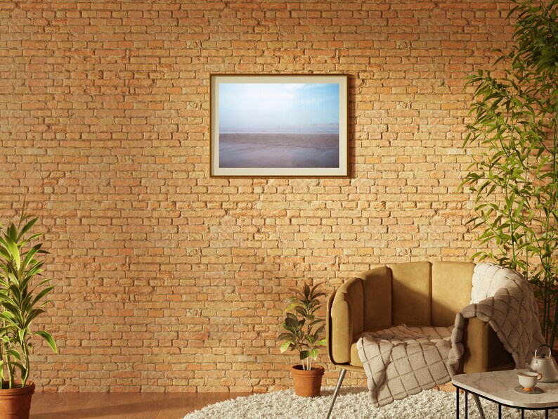 Op de afbeelding: Een bruine leren fauteuil met een wit plaid staat voor een bakstenen muur. Een ingelijst beeld van een strandsc&egrave;ne hangt aan de muur boven de stoel. Een kleine plant in een pot staat op de vloer naast de stoel.