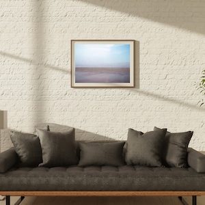 Op de afbeelding: Een grijze bank met een houten frame en donkergrijze kussens staat voor een witte bakstenen muur. Een ingelijste foto van een strandsc&egrave;ne hangt boven de bank. Een hoge gouden staande lamp staat links van de bank. Een plant in een pot staat rechts van de bank.