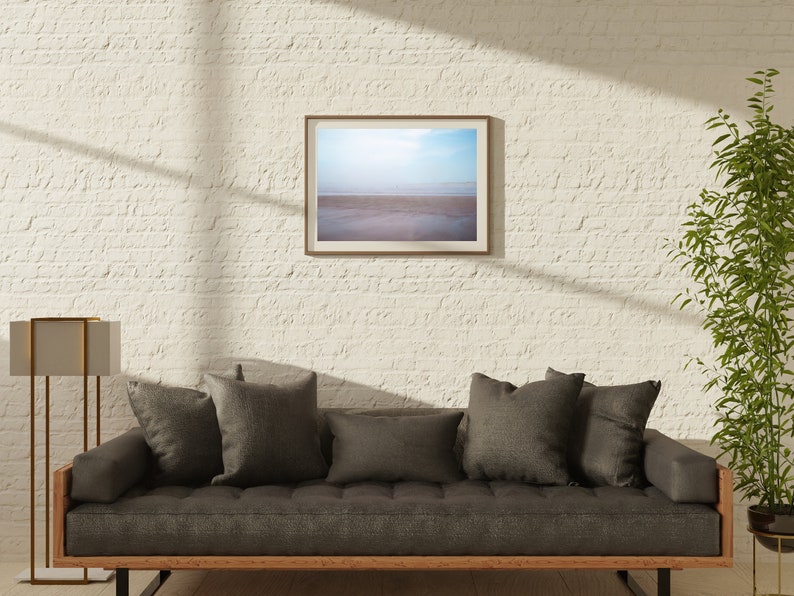 Op de afbeelding: Een grijze bank met een houten frame en donkergrijze kussens staat voor een witte bakstenen muur. Een ingelijste foto van een strandsc&egrave;ne hangt boven de bank. Een hoge gouden staande lamp staat links van de bank. Een plant in een pot staat rechts van de bank.