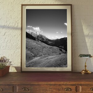 Op de afbeelding: Een zwart-witfoto van een bergketen met een bewolkte lucht. De foto is ingelijst in een bruin houten lijst en staat op een houten tafel met een lamp.