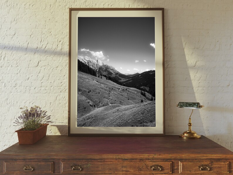Op de afbeelding: Een zwart-witfoto van een bergketen met een bewolkte lucht. De foto is ingelijst in een bruin houten lijst en staat op een houten tafel met een lamp.