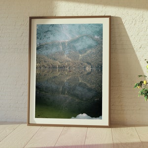 May include: A framed photograph of a mountain lake with a small boat in the middle of the lake. The mountains are reflected in the water. The photo is hanging on a white brick wall.