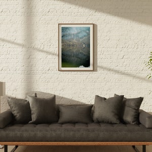 May include: A gray sofa with dark gray throw pillows sits in front of a white brick wall. A framed photograph of a lake with a mountain reflection hangs on the wall. A tall gold floor lamp stands to the left of the sofa. A potted plant is to the right of the sofa.