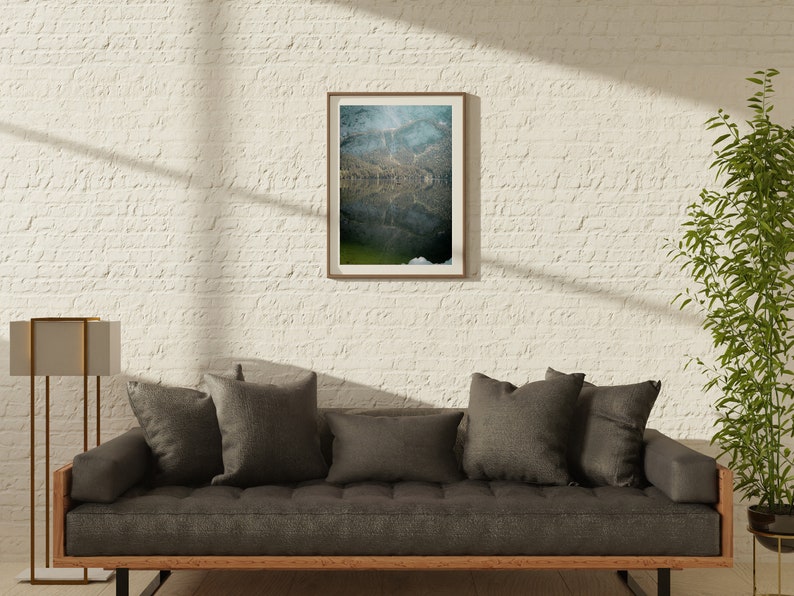 May include: A gray sofa with dark gray throw pillows sits in front of a white brick wall. A framed photograph of a lake with a mountain reflection hangs on the wall. A tall gold floor lamp stands to the left of the sofa. A potted plant is to the right of the sofa.