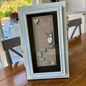 Puede incluir: Obra de arte enmarcada con guijarros que representa una figura sosteniendo un globo en forma de corazón. La obra de arte está sobre un fondo neutro dentro de un marco azul claro. El marco tiene un borde decorativo con cuentas y está apoyado sobre una mesa de madera.