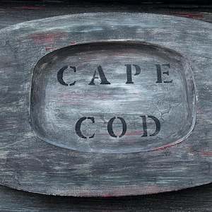May include: A gray wooden sign with the words "CAPE COD" painted in black letters.