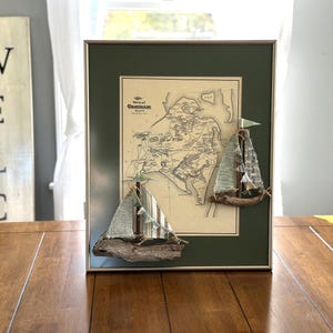 Può includere: Opera d'arte nautica incorniciata con una mappa vintage di Chatham, Mass. Tre barche a vela in miniatura con vele in tessuto e basi in legno galleggiante sono attaccate alla cornice. La cornice ha un passe-partout verde e una cornice in metallo argentato.
