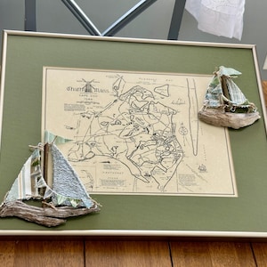 Op de afbeelding: Ingelijste kaart van Chatham, Massachusetts, Cape Cod, met twee kleine, handgemaakte zeilbootdecoraties. De kaart zit in een groene lijst met een witte rand. De zeilboten zijn gemaakt van hout en stof, met blauwe en groene accenten.