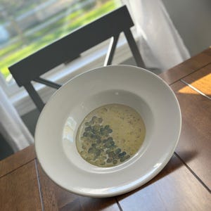 May include: A white ceramic bowl with a shallow depth, containing a light yellow liquid and small, green, four-leaf clover shapes. The bowl sits on a wooden table, with a gray chair and window in the background.