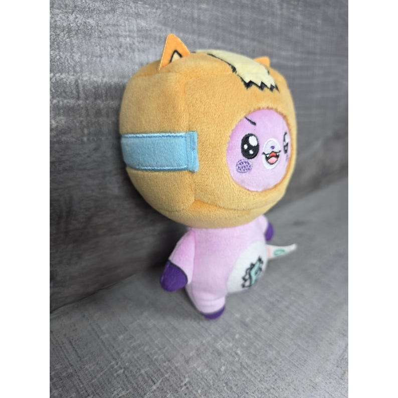 May include: A plush toy featuring a light purple creature wearing a tan, box-shaped head covering with light blue side accents. The creature has large black eyes, a small mouth, and a white belly patch with a small green graphic design.