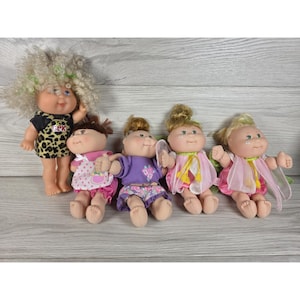 May include: Five dolls with yarn hair and soft bodies. The first doll has blonde curly hair and a leopard print outfit. The others wear various colourful dresses and tops with floral or polka dot patterns, featuring small wings and detailed facial features.
