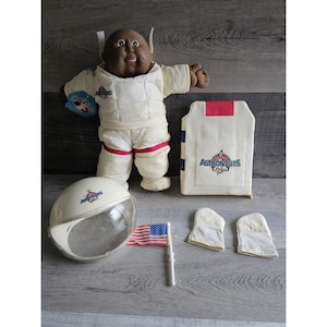 May include: A black Cabbage Patch Kid doll dressed in a white astronaut suit with red accents. The doll is holding a small American flag. The doll is also wearing a white astronaut helmet with a clear visor. The doll is sitting on a wooden surface. The doll is wearing a white astronaut vest with the word "Astronauts" printed on it. The doll is also wearing a pair of white astronaut gloves.