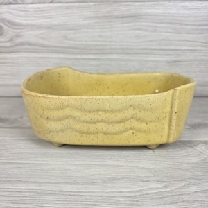 May include: A yellow ceramic bathtub-shaped dish with a speckled finish and embossed wave patterns along the exterior sides. The vessel features four small feet at the base and an open top, designed with a high back edge and a lower front rim.