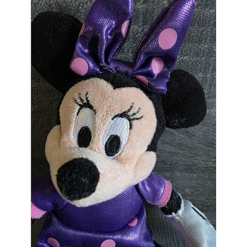 Op de afbeelding: Een pluche Minnie Mouse pop met een zwarte kop en oren, een lichtroze gezicht en een paarse jurk met roze stippen. De pop heeft een paarse strik met roze stippen en zwarte wimpers.