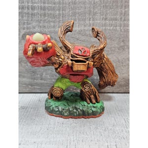 May include: A detailed figurine of a fantasy creature with a tree-like appearance. The figure has a red head and chest, brown wooden arms and horns, and green legs. It stands on a green base with a textured surface.