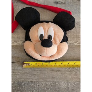 May include: A plush Mickey Mouse face with black ears, a red strap, and a beige face. The face has black eyes and nose, and a smiling mouth. A yellow tape measure is next to the face, showing measurements in inches.