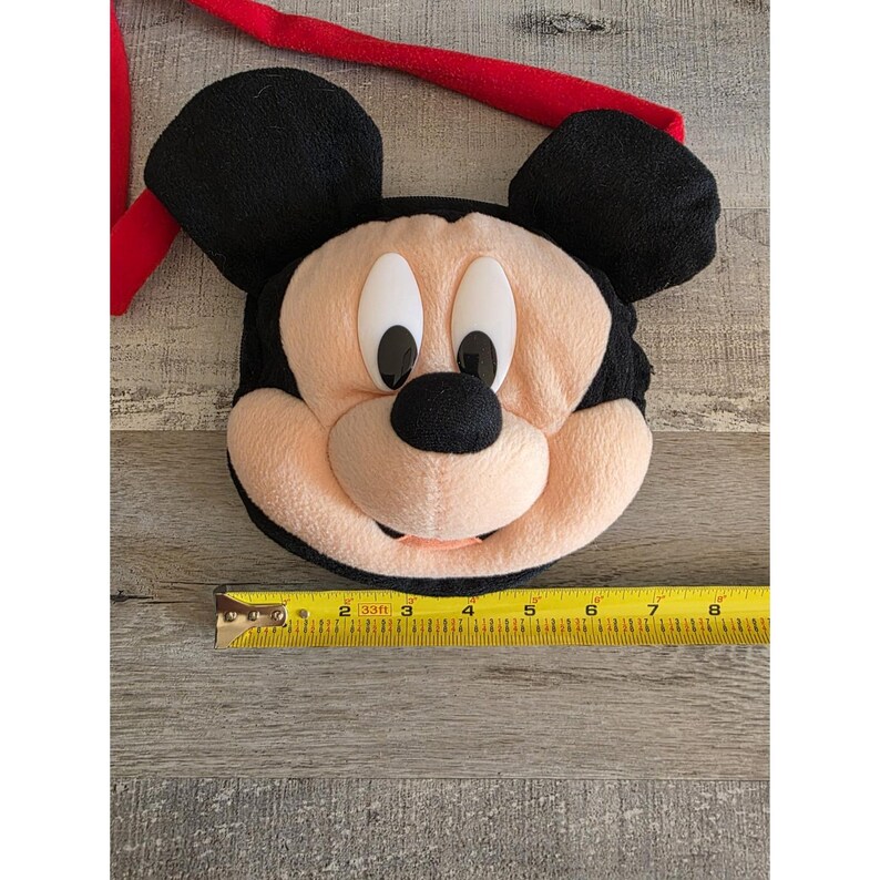 May include: A plush Mickey Mouse face with black ears, a red strap, and a beige face. The face has black eyes and nose, and a smiling mouth. A yellow tape measure is next to the face, showing measurements in inches.