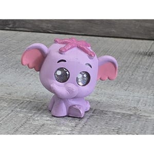 May include: A small, lavender elephant figurine with large, glittery eyes. The elephant has pink inner ears and a pink bow on its head. The figurine is sitting on a gray surface.