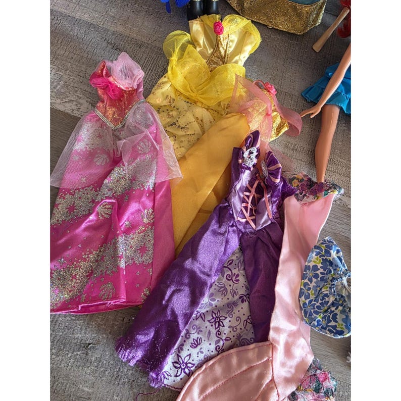 Op de afbeelding: Een verzameling kleurrijke poppenjurken in verschillende stijlen. De jurken omvatten een roze satijnen jurk met zilveren accenten, een gele jurk met een roosdetail en een paarse jurk met bloempatronen. Een blauw bloemenaccessoire is ook zichtbaar.