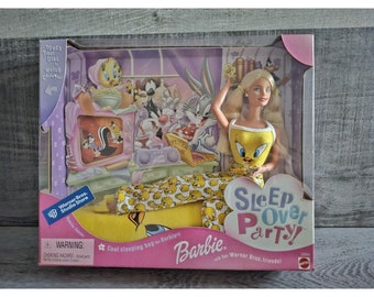 Puede incluir: Muñeca Barbie con un pijama amarillo y blanco con un diseño de Piolín. La muñeca sostiene una bolsa de dormir amarilla y blanca con un diseño de Piolín. El fondo presenta personajes del programa de dibujos animados de Warner Bros., incluyendo Piolín, Silvestre el gato y Bugs Bunny. La caja dice "Sleep Over Party! with her Warner Bros. friends!"