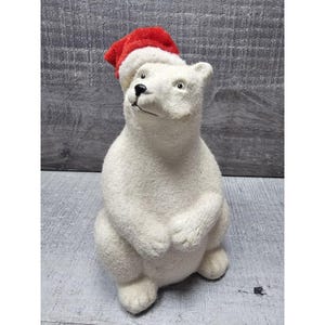May include: A white polar bear ornament wearing a red and white Santa hat. The bear is in a seated position with its paws clasped, gazing upwards. The ornament has a textured finish and is a festive Christmas decoration.