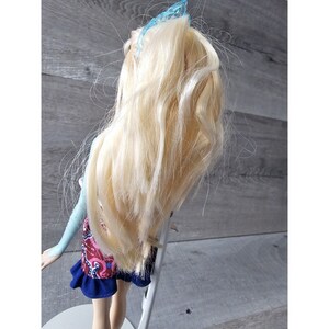 Puede incluir: Una mu&ntilde;eca rubia con una pinza azul en el pelo, que lleva una blusa azul y blanca y una falda con estampado rojo, azul y blanco.