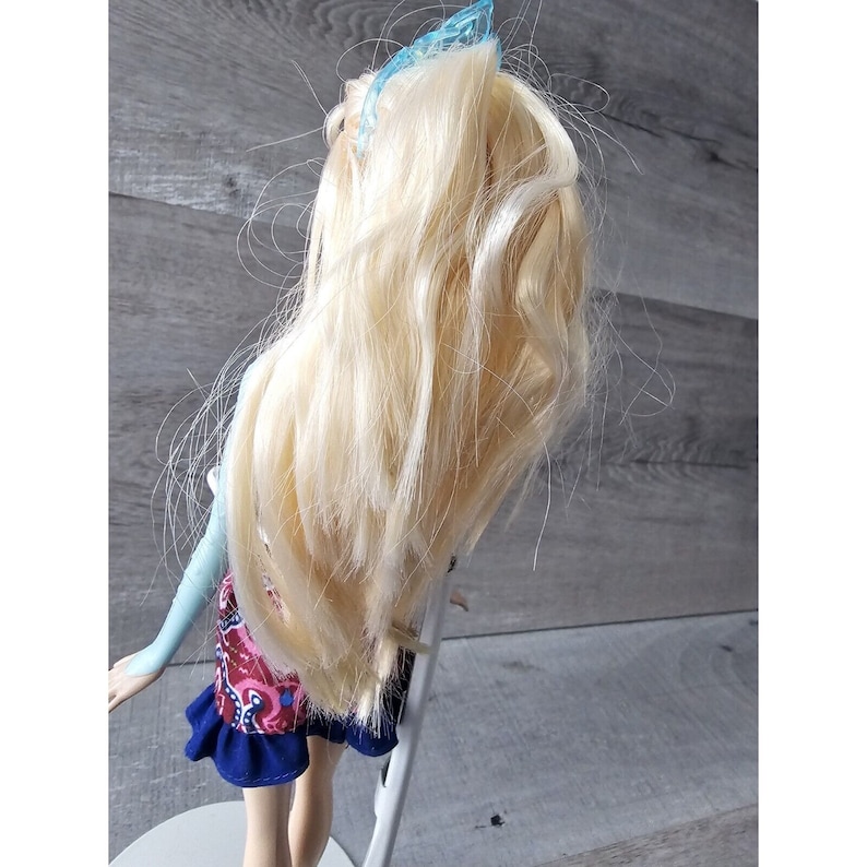 Puede incluir: Una mu&ntilde;eca rubia con una pinza azul en el pelo, que lleva una blusa azul y blanca y una falda con estampado rojo, azul y blanco.