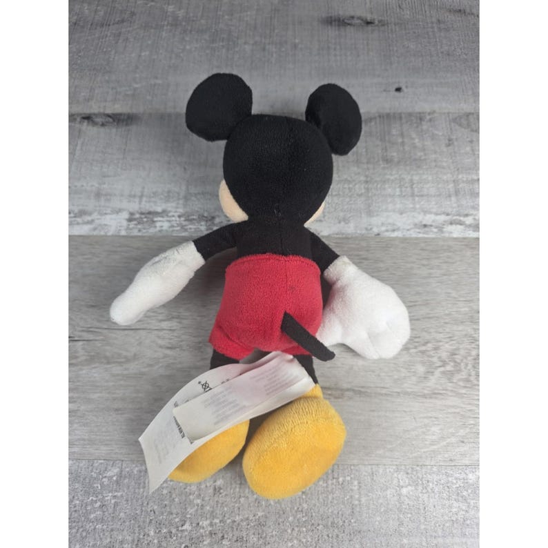 May include: A plush Mickey Mouse toy, viewed from the back, with black ears, a black head, and white gloves. The toy has red shorts, a black tail, and yellow shoes. A white tag is attached.