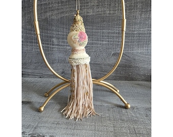 May include: A decorative tassel ornament with a floral design and a long, beige fringe. The ornament is suspended from a gold-colored metal stand. The tassel has a cream-colored beaded trim and a pink floral design.