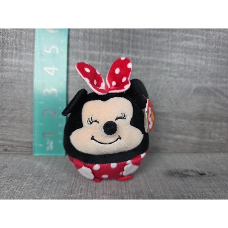 May include: A plush toy of a character with black ears, a cream-coloured face, and a red dress with white polka dots. A red bow with white dots sits on top. A green ruler is visible to the left, showing the toy is approximately 7.6 cm tall.