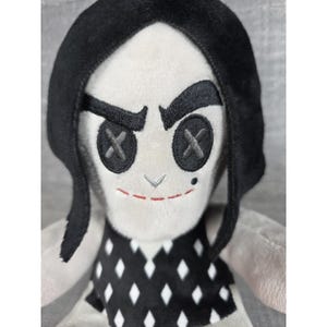 May include: A plush doll with a white face, black hair, and a black and white diamond-patterned dress. The doll has black button eyes with an X design, a stitched red smile, and a small mole.