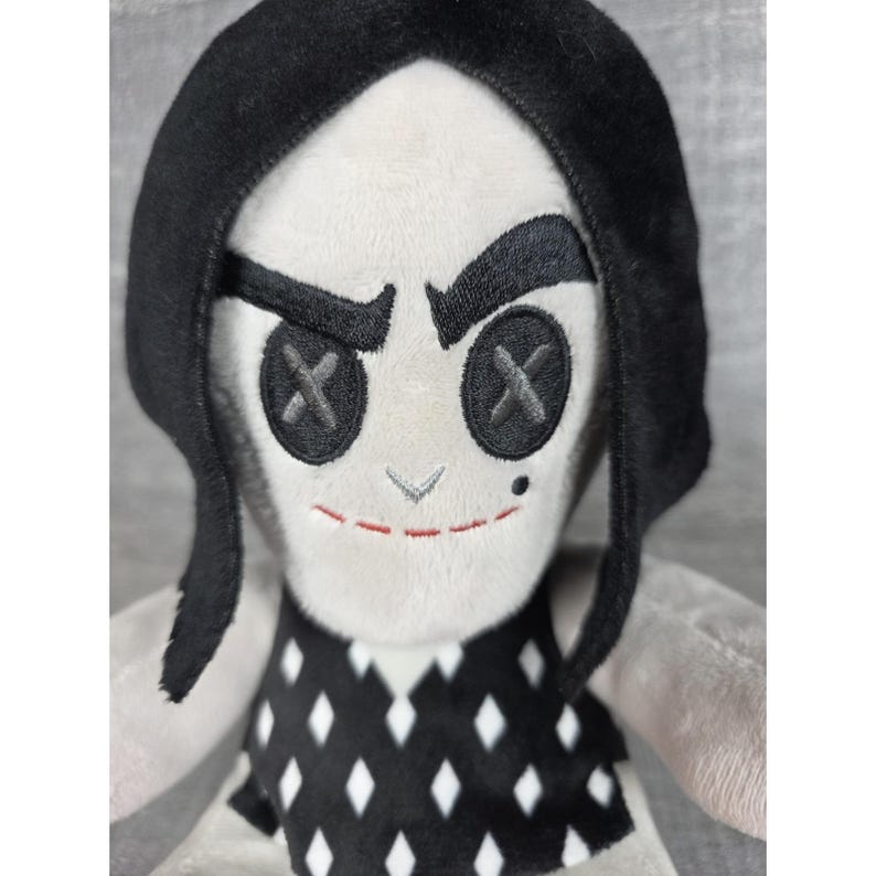 May include: A plush doll with a white face, black hair, and a black and white diamond-patterned dress. The doll has black button eyes with an X design, a stitched red smile, and a small mole.