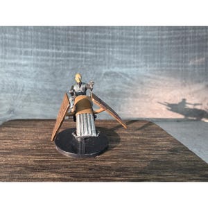 May include: Miniature figure of a person riding a brown and silver vehicle with wing-like structures. The figure is painted with silver and black details. The vehicle sits on a black circular base, with a wooden surface in the foreground.