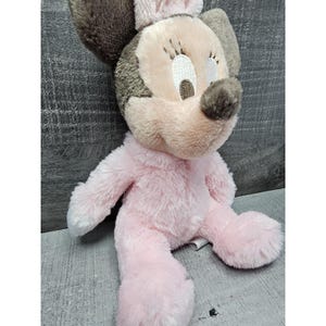 May include: A plush Minnie Mouse doll in a seated position. The doll features a pink body, a light pink face, and brown ears and nose. The doll has closed eyes and a small pink bow on top of its head.