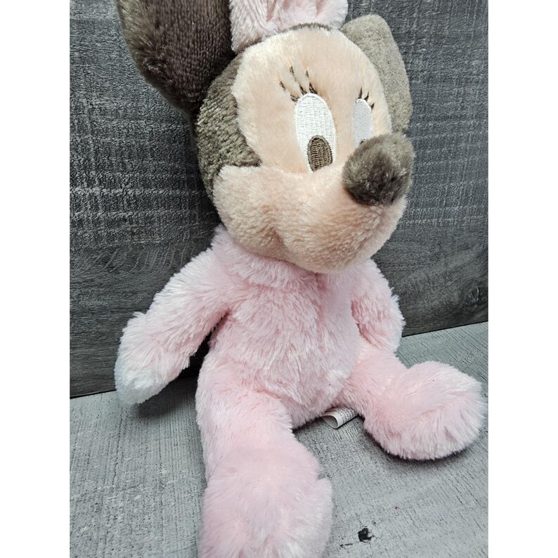 May include: A plush Minnie Mouse doll in a seated position. The doll features a pink body, a light pink face, and brown ears and nose. The doll has closed eyes and a small pink bow on top of its head.