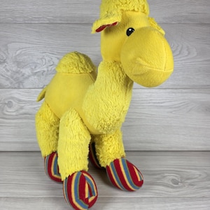 May include: A bright yellow plush camel toy with a soft, textured fabric body. The camel features a single hump on its back, black plastic eyes, and four feet covered in a colorful striped pattern of red, blue, and yellow vertical lines.