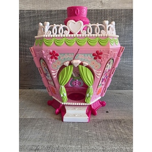 May include: A pink toy playset with the text "Lulu Da Hair & Spa". The playset has a heart-shaped design on top, green curtains, and floral accents. The playset is designed to look like a salon.