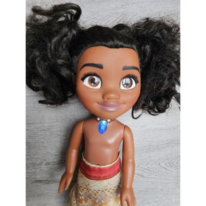 May include: A brown-skinned doll with black curly hair styled in pigtails. The doll is wearing a brown and yellow patterned skirt and a blue necklace with a blue gem pendant.