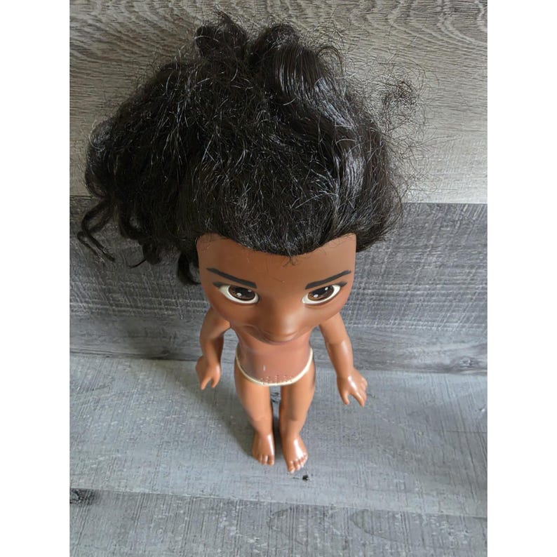 May include: A doll with dark skin, black hair, and a small white garment. The doll has large eyes and is standing on a gray surface. The doll's hair is styled in a messy, voluminous fashion.