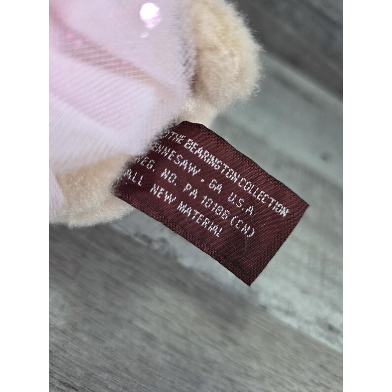 Pu&ograve; includere: Primo piano di un peluche con un'etichetta. L'etichetta &egrave; marrone con testo bianco che recita "THE BEARINGTON COLLECTION ENNESAW - GA U.S.A REG. NO. PA 10186 (CN) ALL NEW MATERIAL". Il giocattolo ha un motivo a quadri rosa e bianco.