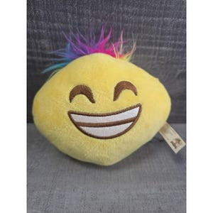 May include: A plush yellow emoji with a smiling face and a colorful mohawk. The emoji has brown eyes and a wide, open mouth with white teeth. A small tag is attached to the side.