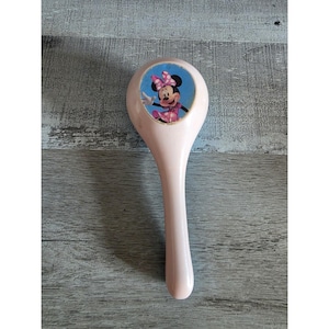 May include: A light pink, vintage-style hairbrush featuring a colorful image of Minnie Mouse. The brush has a long handle and a rounded head with the Minnie Mouse graphic. The brush is likely made of plastic and is designed for children.