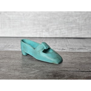 May include: A small, turquoise Mary Jane shoe with a low heel. The vintage-style shoe has a strap across the top and is made of a solid material. The shoe is set against a weathered wooden background.