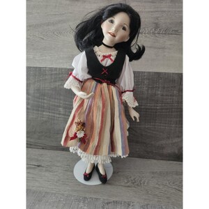May include: A porcelain doll with black hair and a white blouse, black vest, and a colorful striped skirt. The doll is wearing black shoes and is standing on a white base.
