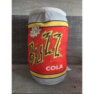 May include: A plush toy shaped like a soda can, featuring a red background with the word "BUZZ" in yellow lightning bolt letters. The can also has the word "COLA" and a "WOW!" speech bubble.