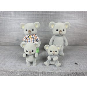May include: Four gray koala figurines. One koala wears a plaid shirt and green pants. The other three koalas are in different poses. The koalas have small black eyes and brown noses. The background is a light gray wooden surface.