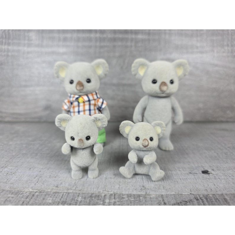 May include: Four gray koala figurines. One koala wears a plaid shirt and green pants. The other three koalas are in different poses. The koalas have small black eyes and brown noses. The background is a light gray wooden surface.