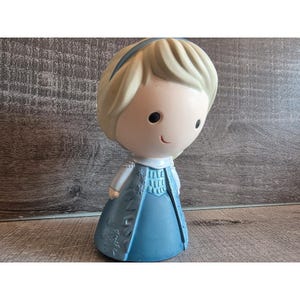 May include: A figurine of a cartoon character with a light blue dress and a blonde hairstyle. The figurine has a small smile and a blue headband. The dress has a detailed design. The figurine is standing on a wooden surface.